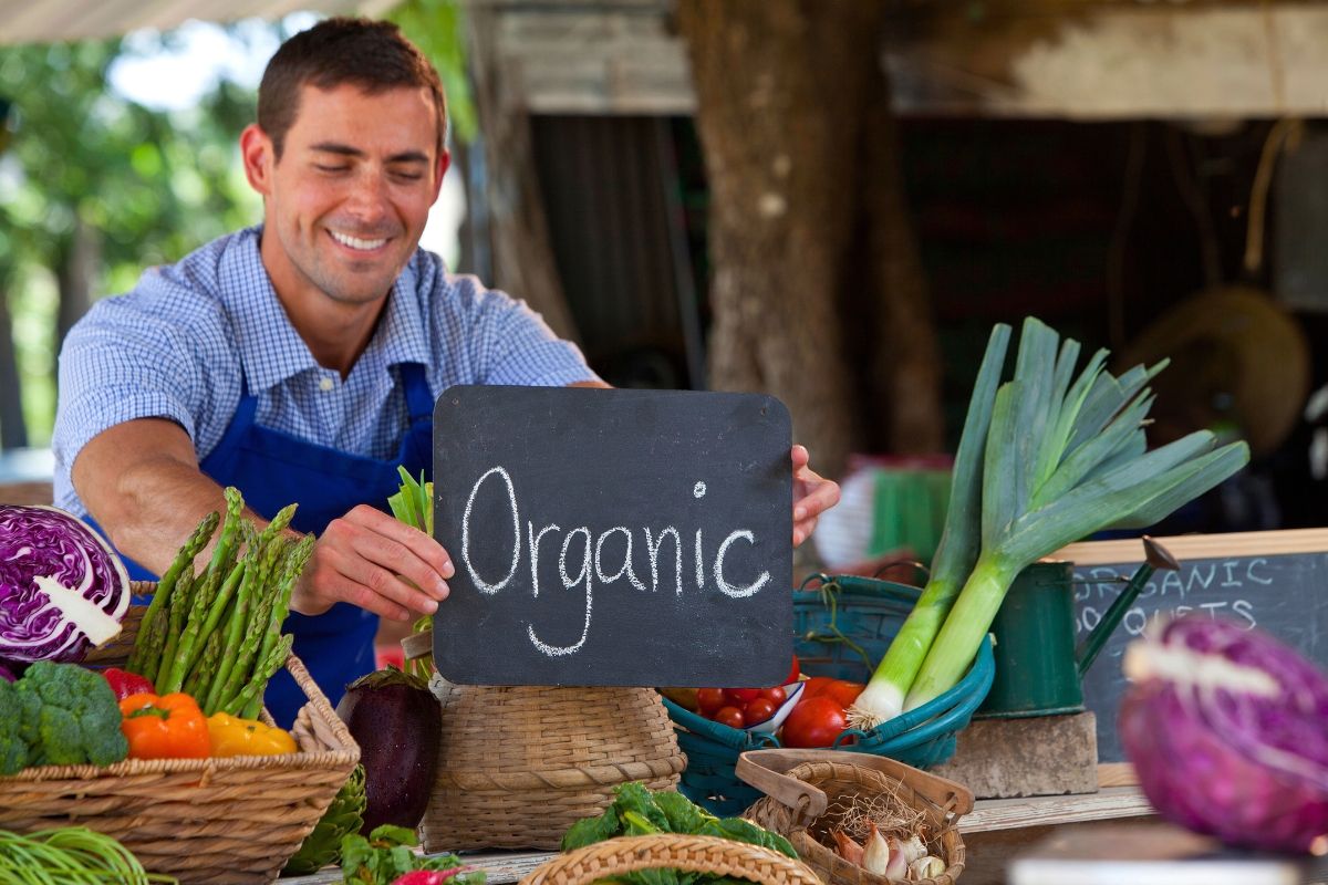Smiling farmers’ market vendor holding a chalkboard “Organic” sign beside baskets of fresh vegetables